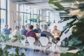 Eine Gruppe von Menschen sitzt um einen großen Tisch in einem modernen Büroraum mit Pflanzen und Deckenbeleuchtung.