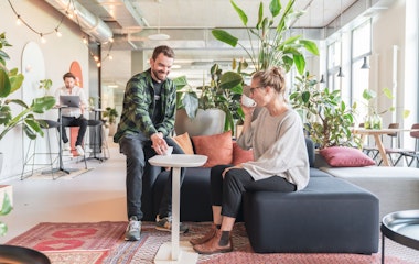 Zwei Personen in einer modernen Bürolounge, umgeben von Pflanzen. Eine Person sitzt auf einem Sofa und trinkt aus einer Tasse, während die andere daneben steht, lächelt und auf einen kleinen Tisch zeigt.