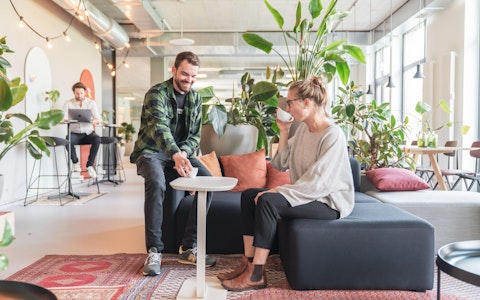 Zwei Personen in einer modernen Bürolounge, umgeben von Pflanzen. Eine Person sitzt auf einem Sofa und trinkt aus einer Tasse, während die andere daneben steht, lächelt und auf einen kleinen Tisch zeigt.