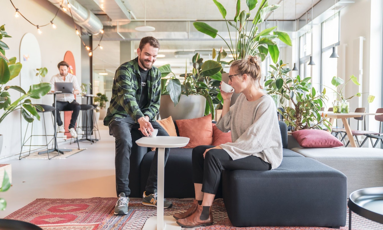 Zwei Personen in einer modernen Bürolounge, umgeben von Pflanzen. Eine Person sitzt auf einem Sofa und trinkt aus einer Tasse, während die andere daneben steht, lächelt und auf einen kleinen Tisch zeigt.