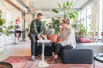 Zwei Personen in einer modernen Bürolounge, umgeben von Pflanzen. Eine Person sitzt auf einem Sofa und trinkt aus einer Tasse, während die andere daneben steht, lächelt und auf einen kleinen Tisch zeigt.