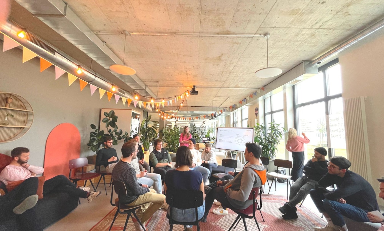 Eine Gruppe von Menschen sitzt im Kreis in einem modernen Raum mit Betondecke, Hängelampen und großen Fenstern. Eine Person steht neben einem Bildschirm, auf dem eine Präsentation läuft.