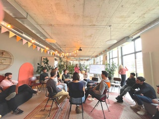 Eine Gruppe von Menschen sitzt im Kreis in einem modernen Raum mit Betondecke, Hängelampen und großen Fenstern. Eine Person steht neben einem Bildschirm, auf dem eine Präsentation läuft.