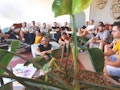 Eine Gruppe von Menschen sitzt und hört in einem Raum zu. Im Vordergrund steht eine Pflanze, auf dem Boden liegen Haftnotizen.