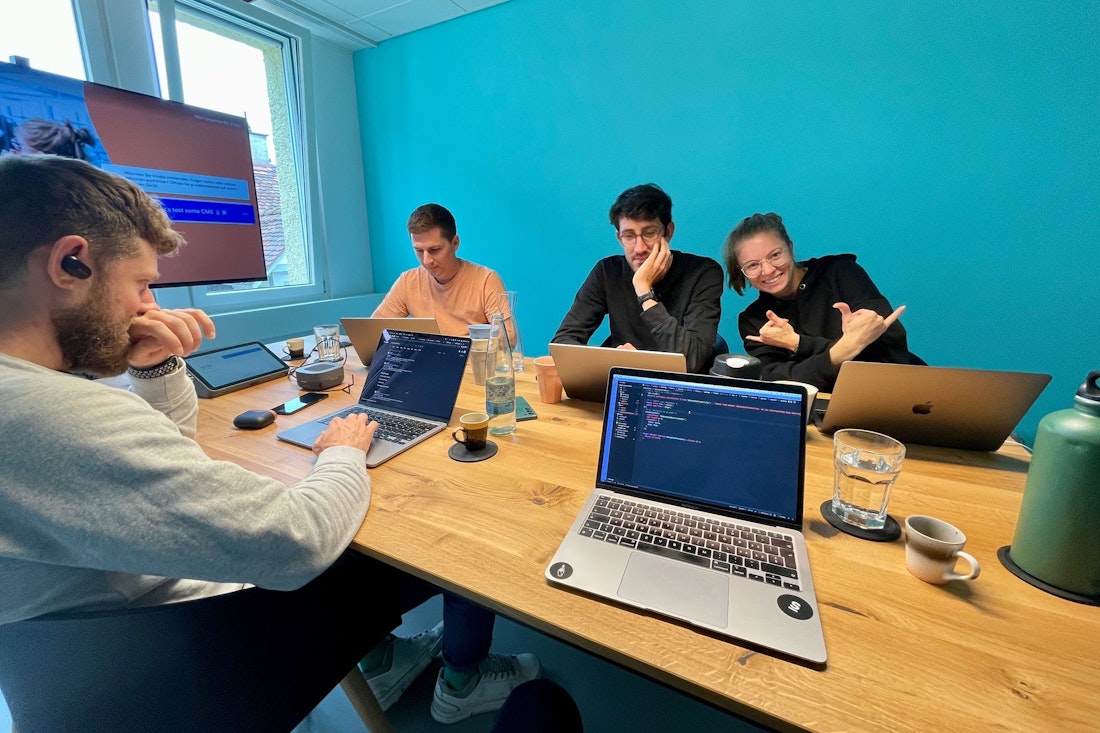 Vier Personen sitzen mit aufgeklappten Laptops an einem Tisch und arbeiten. Eine Person zeigt mit dem Daumen nach oben. An der Wand hinter ihnen hängt ein großer Bildschirm.