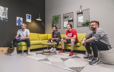 Vier Männer sitzen auf und um ein grünes Sofa herum und spielen Videospiele in einem modernen Wohnzimmer mit Wanddekor und Pflanzen.