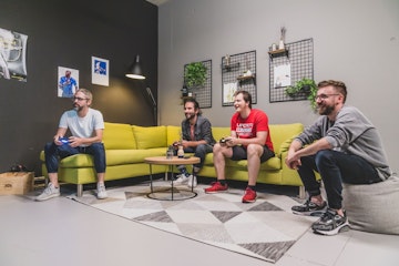 Vier Männer sitzen auf und um ein grünes Sofa herum und spielen Videospiele in einem modernen Wohnzimmer mit Wanddekor und Pflanzen.