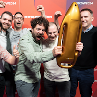 Sieben Männer lächeln und jubeln gemeinsam, während einer von ihnen eine raketenförmige goldene Trophäe hält, die vor einem roten Hintergrund mit Event- und Sponsorenlogo steht.