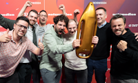 Sieben Männer lächeln und jubeln gemeinsam, während einer von ihnen eine raketenförmige goldene Trophäe hält, die vor einem roten Hintergrund mit Event- und Sponsorenlogo steht.