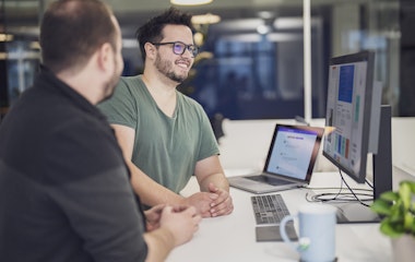 Zwei Personen sitzen an einem Schreibtisch mit einem Laptop und einem großen Monitor und diskutieren über ein auf dem Bildschirm angezeigtes Projekt. Auf dem Schreibtisch stehen außerdem eine Tasse und eine Pflanze.