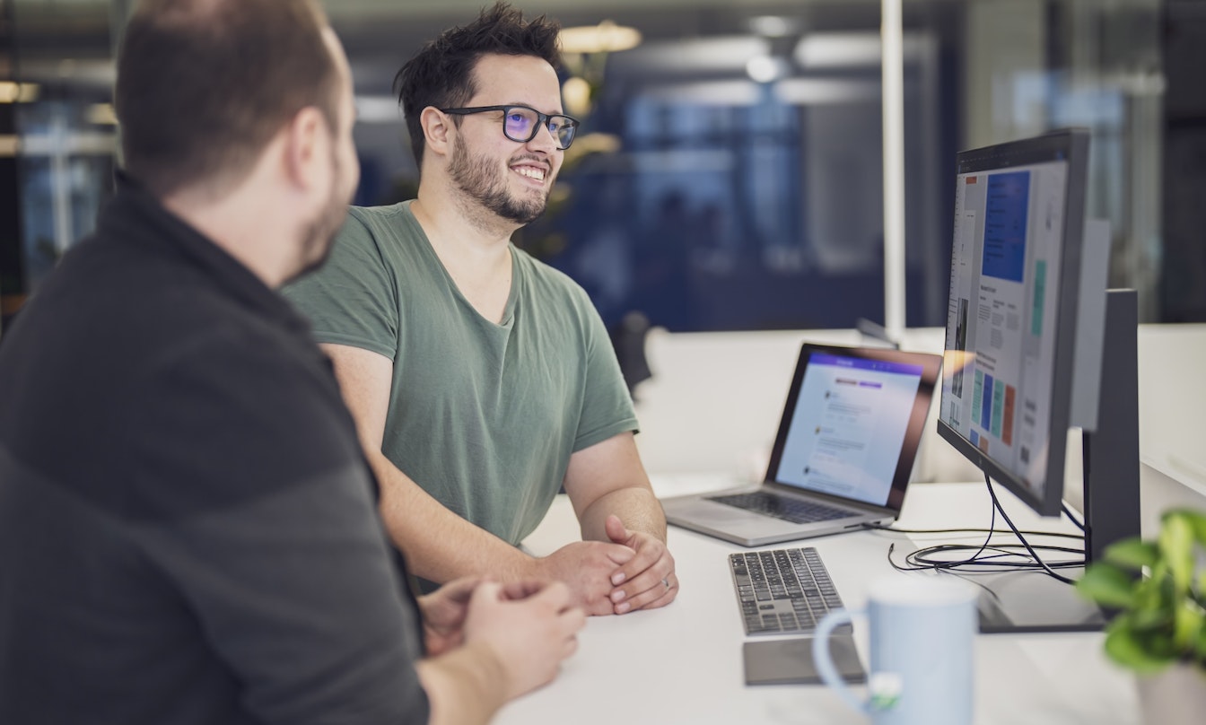 Zwei Personen sitzen an einem Schreibtisch mit einem Laptop und einem großen Monitor und diskutieren über ein auf dem Bildschirm angezeigtes Projekt. Auf dem Schreibtisch stehen außerdem eine Tasse und eine Pflanze.
