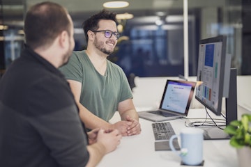 Zwei Personen sitzen an einem Schreibtisch mit einem Laptop und einem großen Monitor und diskutieren über ein auf dem Bildschirm angezeigtes Projekt. Auf dem Schreibtisch stehen außerdem eine Tasse und eine Pflanze.