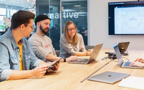 Drei Personen sitzen mit Laptops um einen Konferenztisch und diskutieren. Auf einem an der Wand montierten Bildschirm ist eine Präsentation zu sehen.