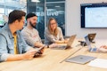 Drei Personen sitzen mit Laptops um einen Konferenztisch und diskutieren. Auf einem an der Wand montierten Bildschirm ist eine Präsentation zu sehen.