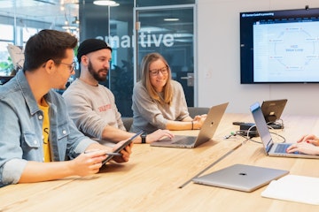 Drei Personen sitzen mit Laptops um einen Konferenztisch und diskutieren. Auf einem an der Wand montierten Bildschirm ist eine Präsentation zu sehen.