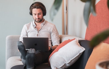 Eine Person mit Kopfhörern sitzt mit einem Laptop auf einer Couch, umgeben von Kissen und Pflanzen.