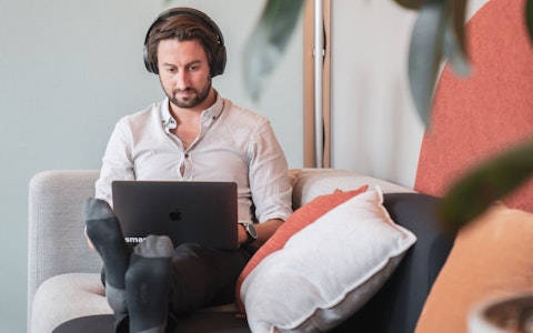 Eine Person mit Kopfhörern sitzt mit einem Laptop auf einer Couch, umgeben von Kissen und Pflanzen.