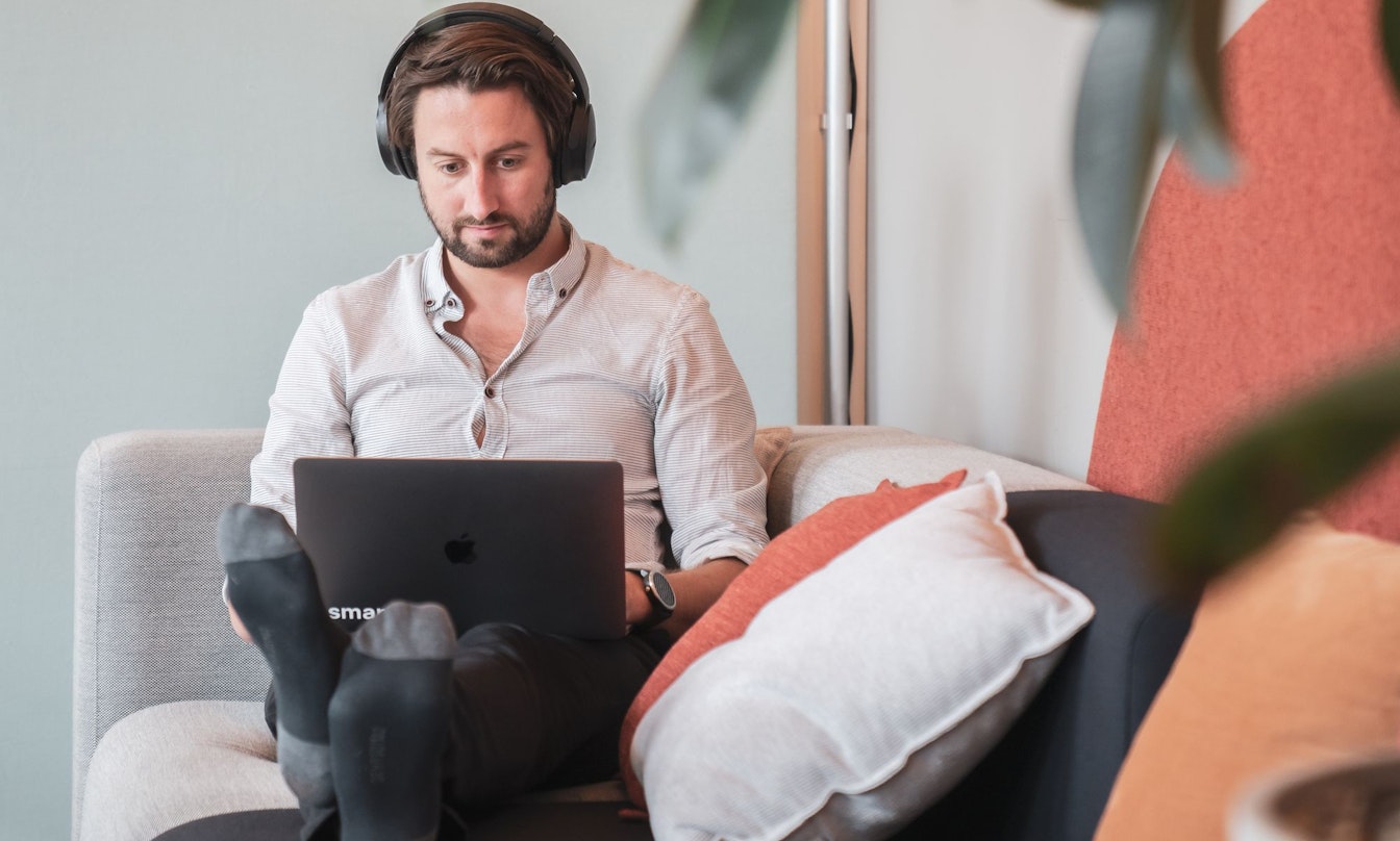 Eine Person mit Kopfhörern sitzt mit einem Laptop auf einer Couch, umgeben von Kissen und Pflanzen.
