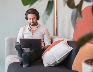 Eine Person mit Kopfhörern sitzt mit einem Laptop auf einer Couch, umgeben von Kissen und Pflanzen.