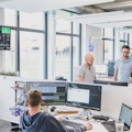 Ein modernes Büro mit vier Personen an Schreibtischen, großen Fenstern und einem Bildschirm mit Diagrammen an der Wand.