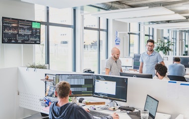 Ein modernes Büro mit vier Personen an Schreibtischen, großen Fenstern und einem Bildschirm mit Diagrammen an der Wand.