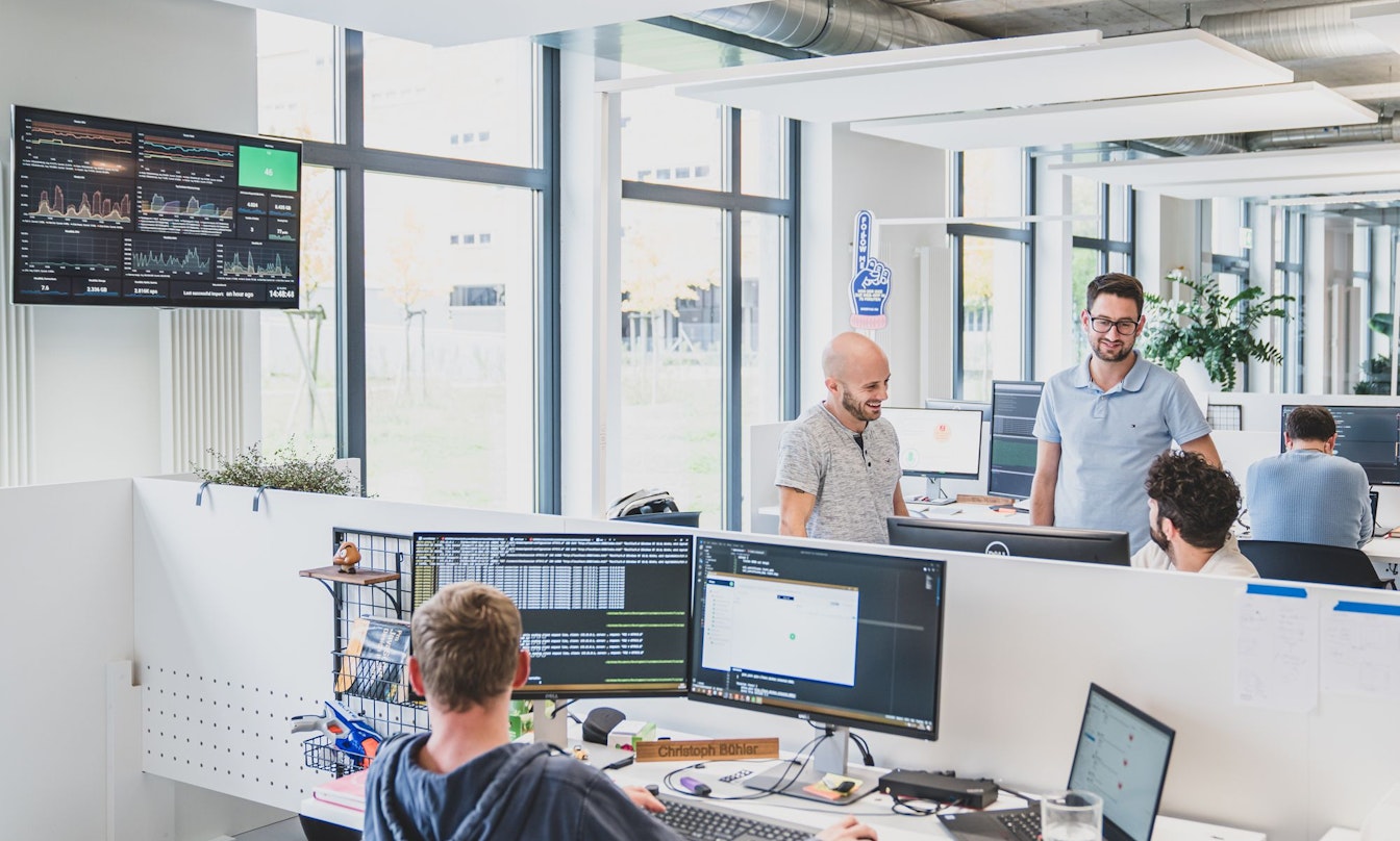 Ein modernes Büro mit vier Personen an Schreibtischen, großen Fenstern und einem Bildschirm mit Diagrammen an der Wand.