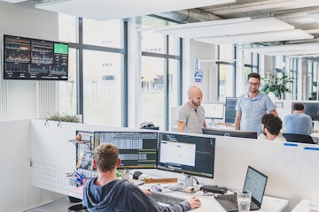 Ein modernes Büro mit vier Personen an Schreibtischen, großen Fenstern und einem Bildschirm mit Diagrammen an der Wand.
