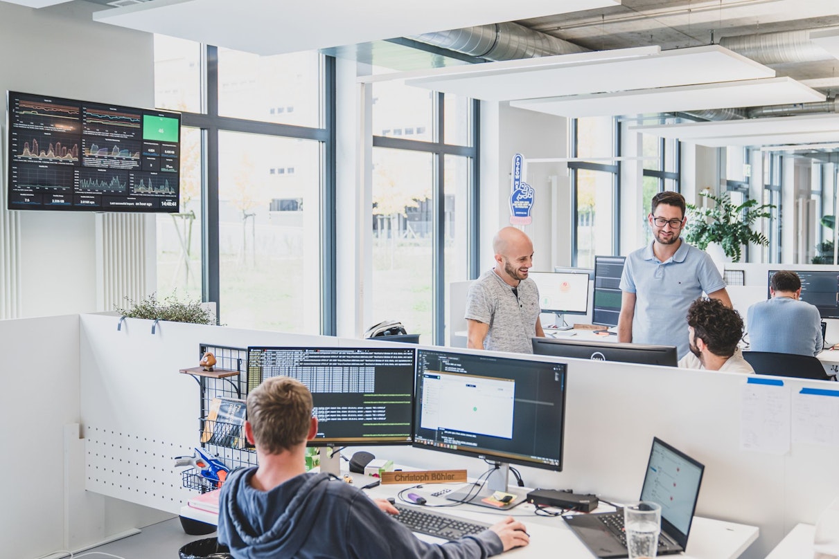 Ein modernes Büro mit vier Personen an Schreibtischen, großen Fenstern und einem Bildschirm mit Diagrammen an der Wand.