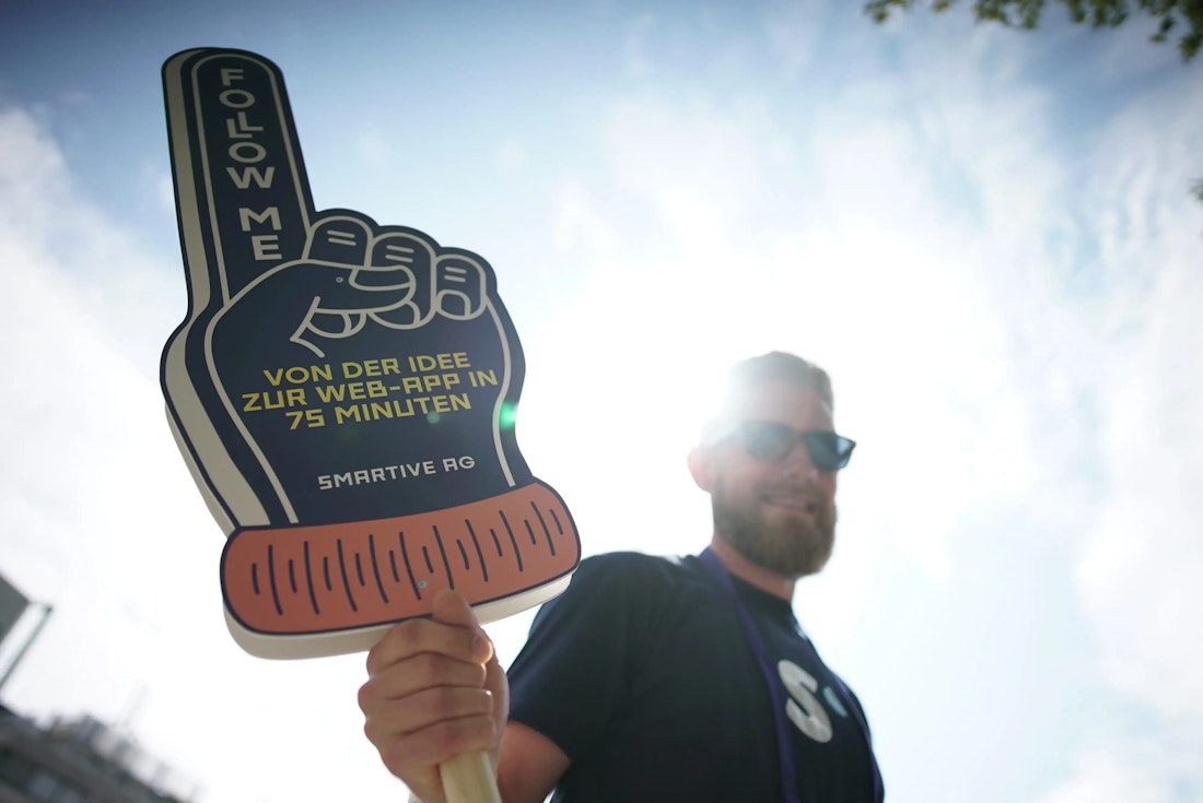 Eine Person hält unter einem strahlend sonnigen Himmel ein großes Schaumstoff-Fingerschild mit der Aufschrift „FOLGEN SIE MIR VON DER IDEE ZUR WEB-APP IN 75 MINUTEN“.