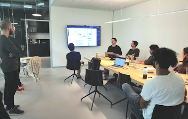 In einem Konferenzraum sitzt eine Gruppe von Menschen an einem langen Tisch und sieht sich eine Präsentation auf einem an der Wand montierten Bildschirm an. Auf dem Tisch sind Laptops und Notizen zu sehen.