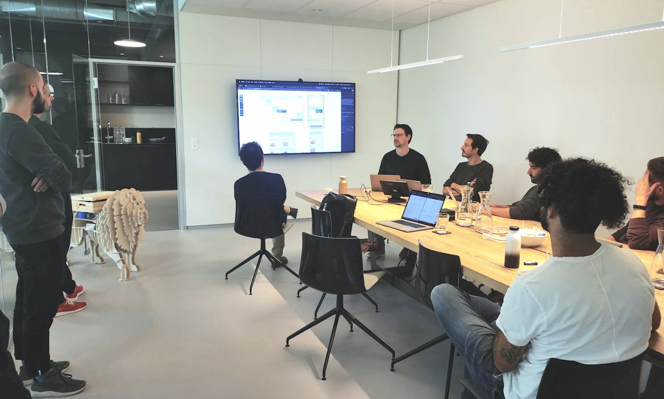 In einem Konferenzraum sitzt eine Gruppe von Menschen an einem langen Tisch und sieht sich eine Präsentation auf einem an der Wand montierten Bildschirm an. Auf dem Tisch sind Laptops und Notizen zu sehen.