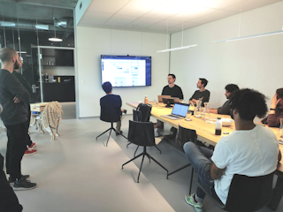 In einem Konferenzraum sitzt eine Gruppe von Menschen an einem langen Tisch und sieht sich eine Präsentation auf einem an der Wand montierten Bildschirm an. Auf dem Tisch sind Laptops und Notizen zu sehen.