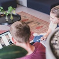 Zwei Personen sitzen auf dem Boden und arbeiten an einem Laptop und einem Tablet in einer gemütlichen Umgebung mit Pflanzen und einem Teppich.