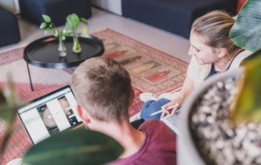Zwei Personen sitzen auf dem Boden und arbeiten an einem Laptop und einem Tablet in einer gemütlichen Umgebung mit Pflanzen und einem Teppich.