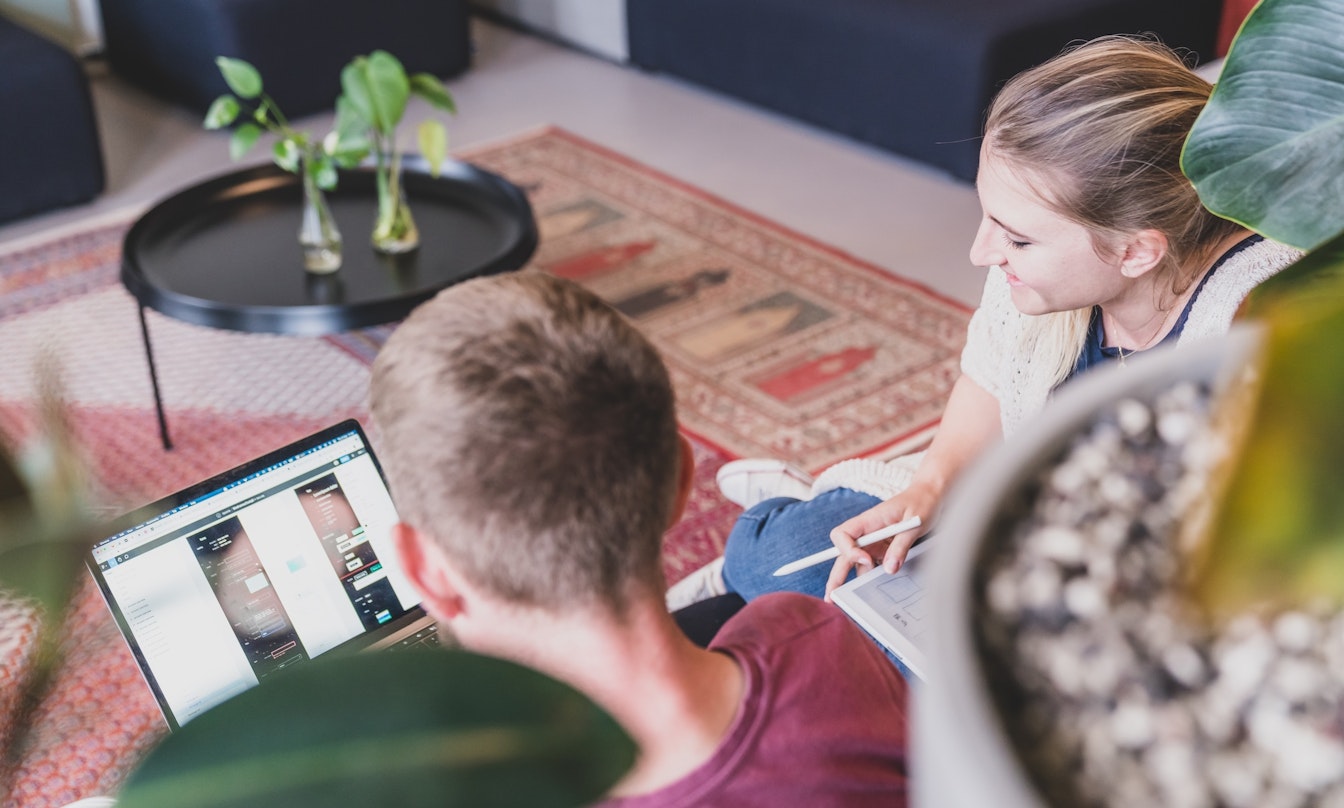 Zwei Personen sitzen auf dem Boden und arbeiten an einem Laptop und einem Tablet in einer gemütlichen Umgebung mit Pflanzen und einem Teppich.
