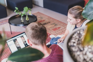 Zwei Personen sitzen auf dem Boden und arbeiten an einem Laptop und einem Tablet in einer gemütlichen Umgebung mit Pflanzen und einem Teppich.