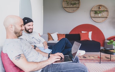 Zwei Männer sitzen auf einem Sofa. Einer benutzt einen Laptop, der andere hält ein Tablet. Der Raum ist modern eingerichtet, mit runden Wandregalen und einem gemusterten Teppich.