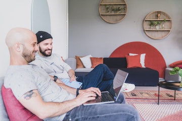 Zwei Männer sitzen auf einem Sofa. Einer benutzt einen Laptop, der andere hält ein Tablet. Der Raum ist modern eingerichtet, mit runden Wandregalen und einem gemusterten Teppich.