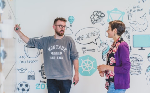 Zwei Personen diskutieren an einem Whiteboard mit illustrierten Grafiken, darunter ein Einhorn und eine Teekanne. Eine Person schreibt, beide sind leger gekleidet.