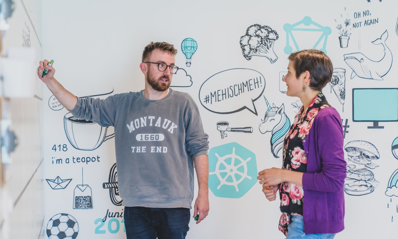 Zwei Personen diskutieren an einem Whiteboard mit illustrierten Grafiken, darunter ein Einhorn und eine Teekanne. Eine Person schreibt, beide sind leger gekleidet.