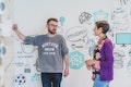 Zwei Personen diskutieren an einem Whiteboard mit illustrierten Grafiken, darunter ein Einhorn und eine Teekanne. Eine Person schreibt, beide sind leger gekleidet.