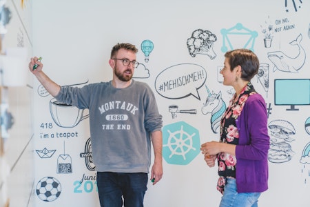 Zwei Personen diskutieren an einem Whiteboard mit illustrierten Grafiken, darunter ein Einhorn und eine Teekanne. Eine Person schreibt, beide sind leger gekleidet.
