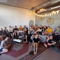 Eine Gruppe von Menschen sitzt auf Sofas und dem Boden in einem modernen, gemĂŒtlichen Raum mit Teppichen und Pflanzen.