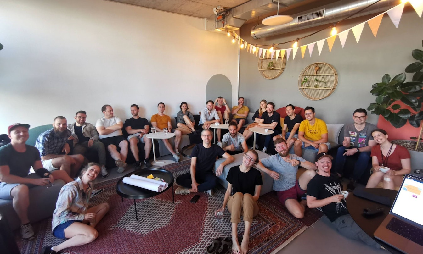 Eine Gruppe von Menschen sitzt auf Sofas und dem Boden in einem modernen, gemütlichen Raum mit Teppichen und Pflanzen.