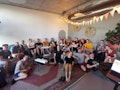 Eine Gruppe von Menschen sitzt auf Sofas und dem Boden in einem modernen, gemütlichen Raum mit Teppichen und Pflanzen.
