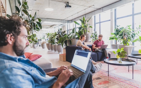 Drei Personen arbeiten an Laptops in einem hellen, modernen Büroraum mit großen Fenstern und viel Grün.