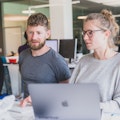 Ein Mann und eine Frau arbeiten gemeinsam an einem Schreibtisch mit Computern in einer modernen Büroumgebung.