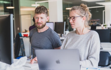 Ein Mann und eine Frau arbeiten gemeinsam an einem Schreibtisch mit Computern in einer modernen Büroumgebung.