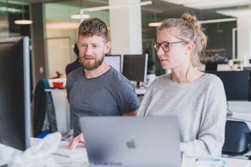 Ein Mann und eine Frau arbeiten gemeinsam an einem Schreibtisch mit Computern in einer modernen Büroumgebung.