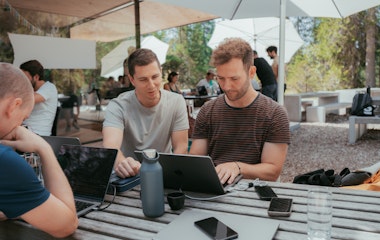 Zwei Personen unter einem grossen weissen Sonnenschrim schauen auf ein Macbook. Gegenüber sitzt eine weitere Person, die am Laptop arbeitet. Ein Smartphone liegt auf dem Tisch.